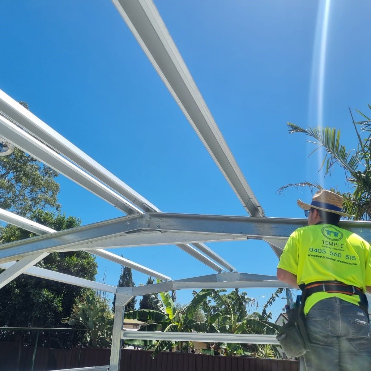 Looking up at a galvanised steel roof frame against clear sky, with a Temple Construction Group tradesperson in branded hi-vis working beneath it.