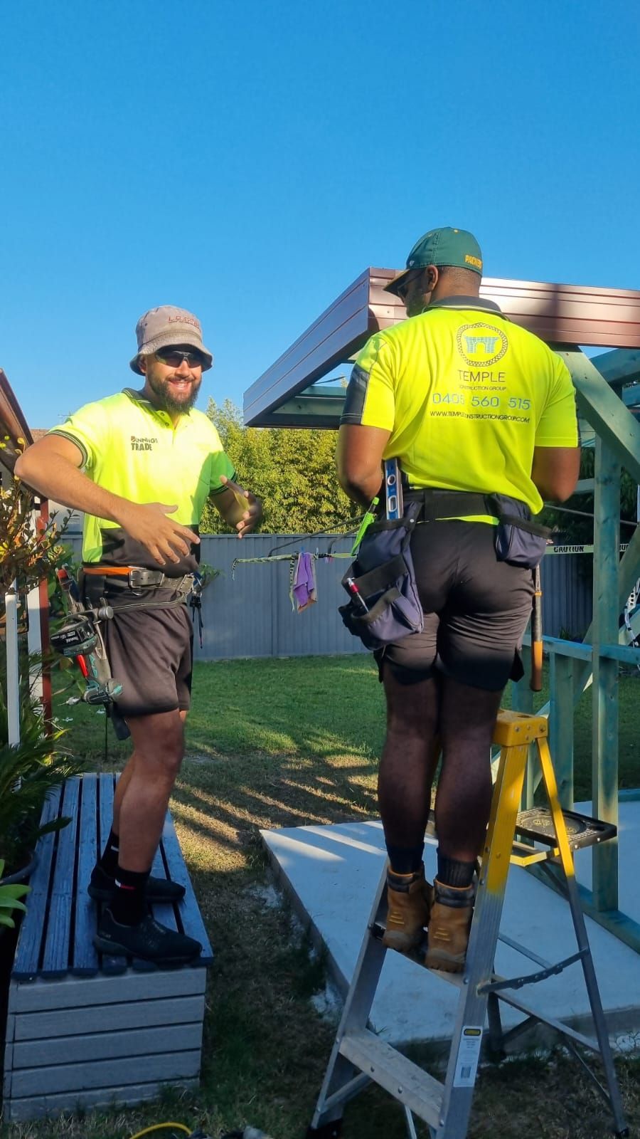 Two tradespeople in hi-vis on a residential job, one on a stepladder under a steel patio frame, tool belts on.