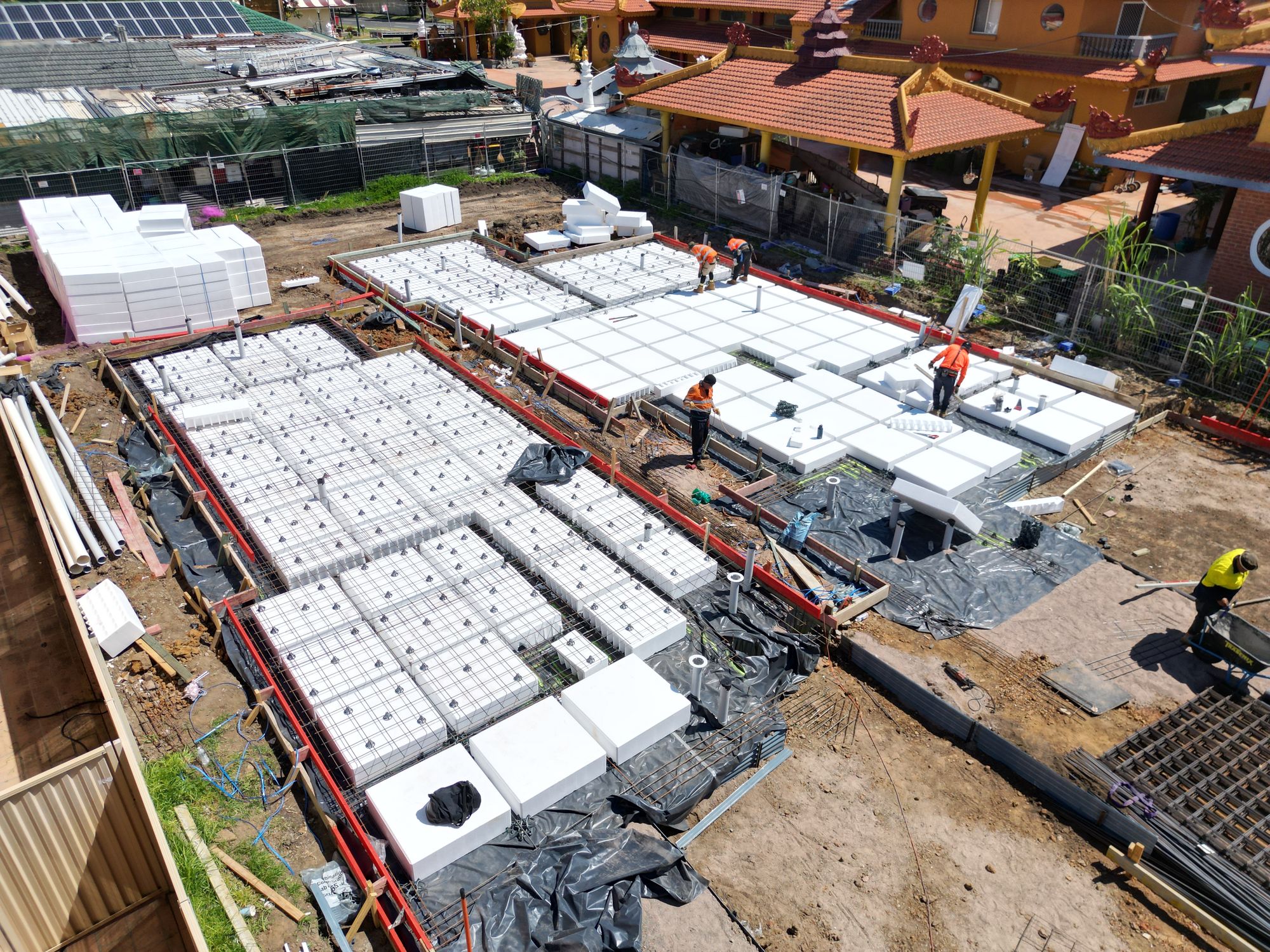 Waffle-pod raft slab laid out: polystyrene void formers on membrane, reinforcement mesh over the top, plumbing risers set, crew placing pods.